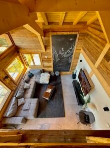 Le salon du Chalet Spencer est un véritable cocon de confort et d’élégance. Avec ses plafonds cathédrale, son immense baie vitrée offrant une vue panoramique sur les montagnes, et son architecture en bois naturel, il invite à la détente et à l’évasion. Son grand canapé d’angle en tissu, agrémenté de coussins moelleux, est l’endroit idéal pour se retrouver en famille ou entre amis. Face à une cheminée moderne et un écran plat, cet espace associe chaleur et modernité pour créer une atmosphère cosy et raffinée. Le mélange subtil de matériaux nobles et de touches contemporaines, comme l’illustration murale rappelant les sommets alpins, fait de ce salon un lieu unique où chaque instant devient inoubliable.